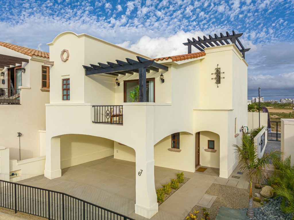 Cream stucco, two‑story coastal villa with arched carport, black wooden pergola, and a balcony overlooking a paved driveway under a blue sky.