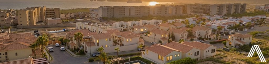 Coastal suburban neighborhood at sunset with rows of beige, tile-roof houses and palm trees, and mid-rise buildings along the shore; the ocean is visible on the horizon.